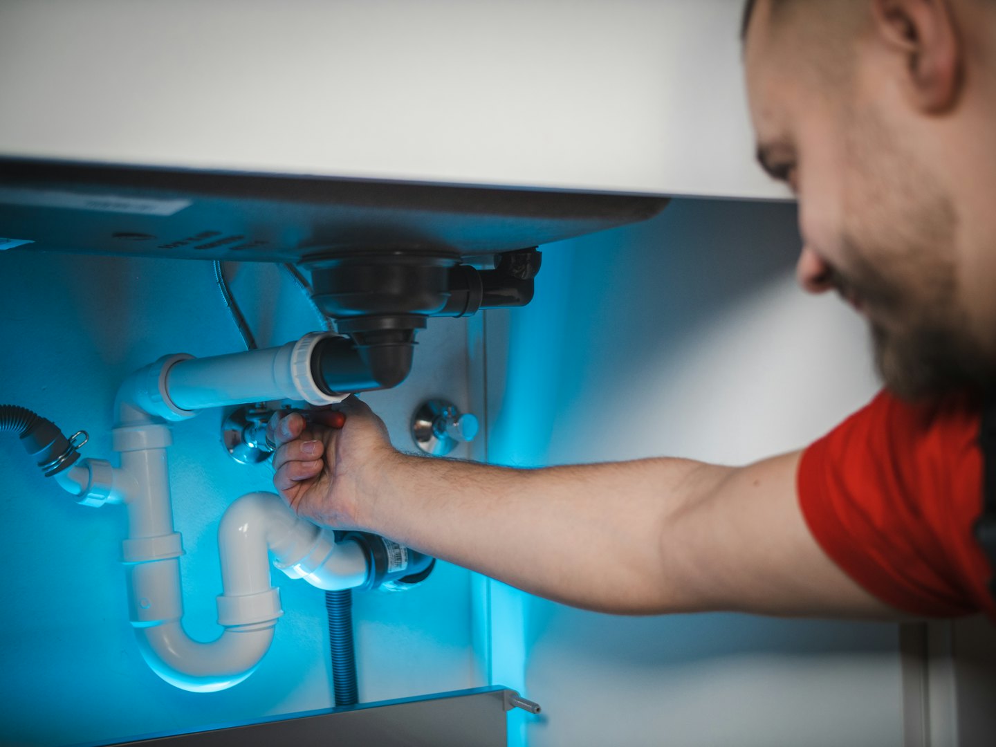 Man in red shirt works on pipes under a sink.